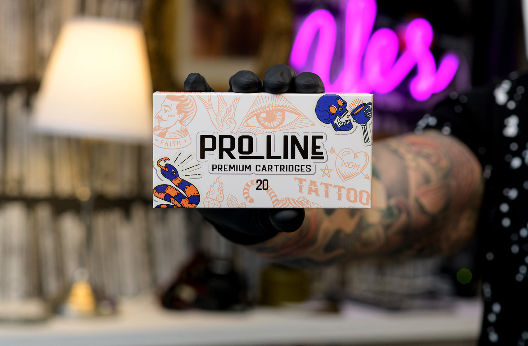 Photograph of a hand wearing black gloves holding a box filled with Pro Line Cartridges brand cartridges. The box is positioned in the center of the photo, with the cartridges visible inside. The background behind the box is blurred, emphasizing the focus on the product being held.