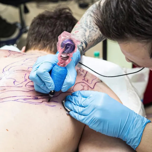 A tattoo artist working on a large back tattoo design. The artist is using a tattoo machine with a pink plastic cover and wearing blue gloves. The client lies face down, and the outline of the tattoo is visible on their back.