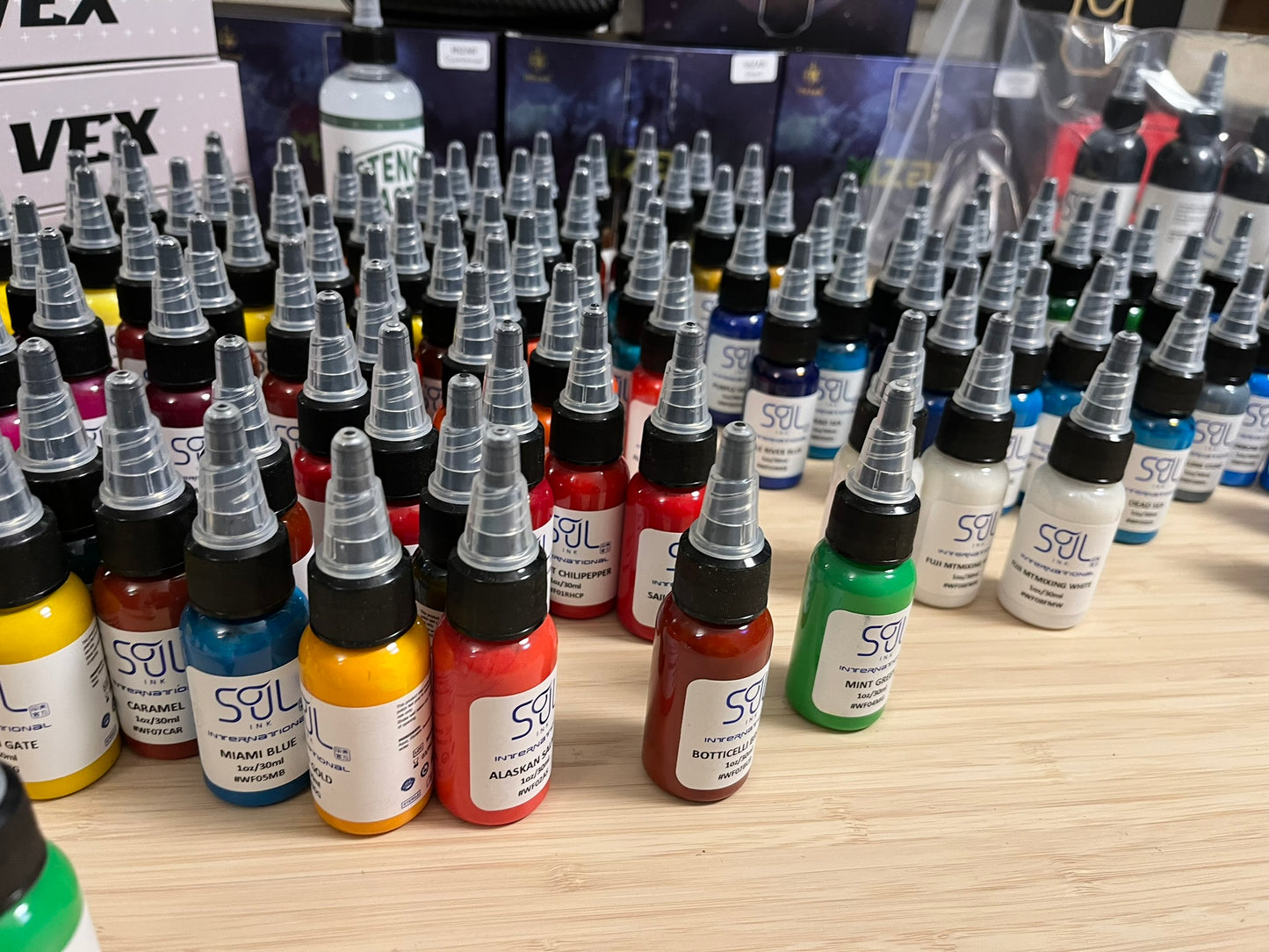 Bottles of 'Soul' brand ink in various colors are neatly arranged on a table. Each bottle has a label with the color name and a hexadecimal code, such as 'Miami Blue' (#WF05MB), 'Alaskan Salmon' (#WF02AS), and 'Botticelli Brown' (#WF07BCB). The bottles are equipped with dispensing tips, and multiple colors can be seen including red, blue, yellow, green, and white.