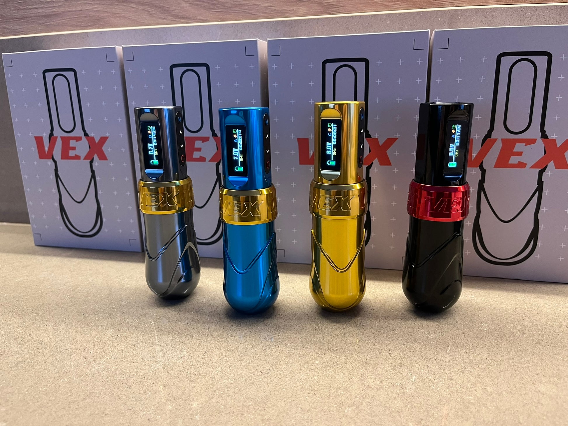 Four Vex tattoo machines in different colors: gold, black and red, blue and gold, and grey and gold. Behind each machine is its corresponding packaging box